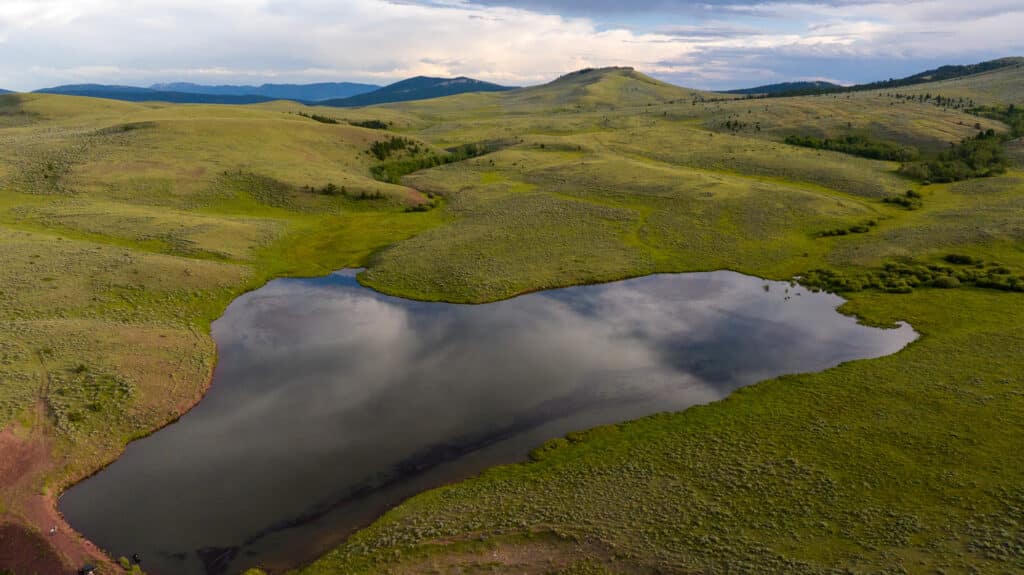 A small, heart-shaped lake reflects the cloudy sky, surrounded by rolling green hills and distant mountains under a blue and cloudy sky at Battle Mountain Ranch.