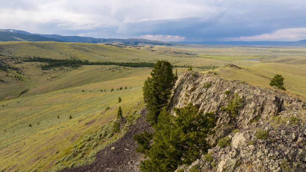 A rocky ridge with scattered trees overlooks the vast, rolling green plain of Battle Mountain Ranch under a partly cloudy sky, with distant hills and mountains on the horizon.
