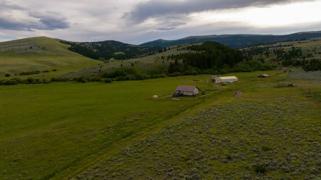 Battle Mountain Ranch, a rural ranch with several buildings, sits in a green valley surrounded by rolling hills and forests under a cloudy sky.
