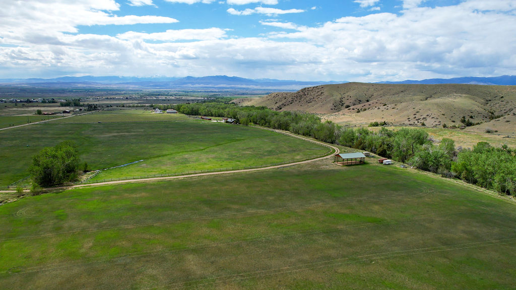 Aerial view of a large, green rural landscape with scattered trees, a winding dirt road, hills, and distant mountains under a partly cloudy sky—featuring the picturesque expanse of a horse ranch alongside the gentle flow of Wisconsin Creek.
