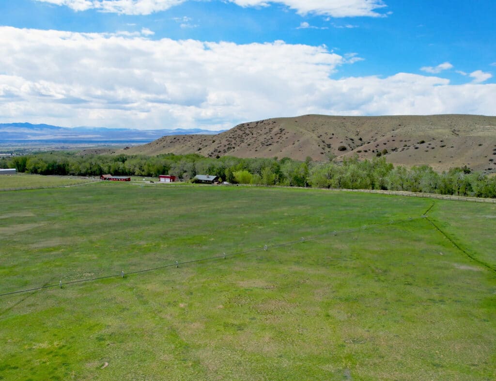 Wide open grassy field with scattered trees, a few farm buildings in the distance, and rolling hills under a blue sky with clouds, reminiscent of a peaceful horse ranch near Wisconsin Creek.
