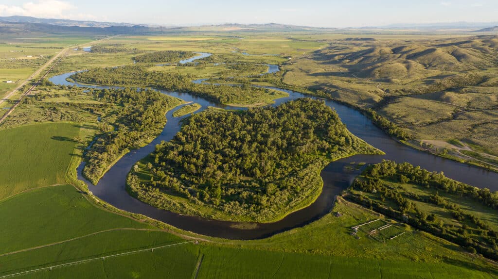 A winding river flows through green fields and forested areas, where cattle farming thrives, with mountains visible in the distance under a partly cloudy sky.