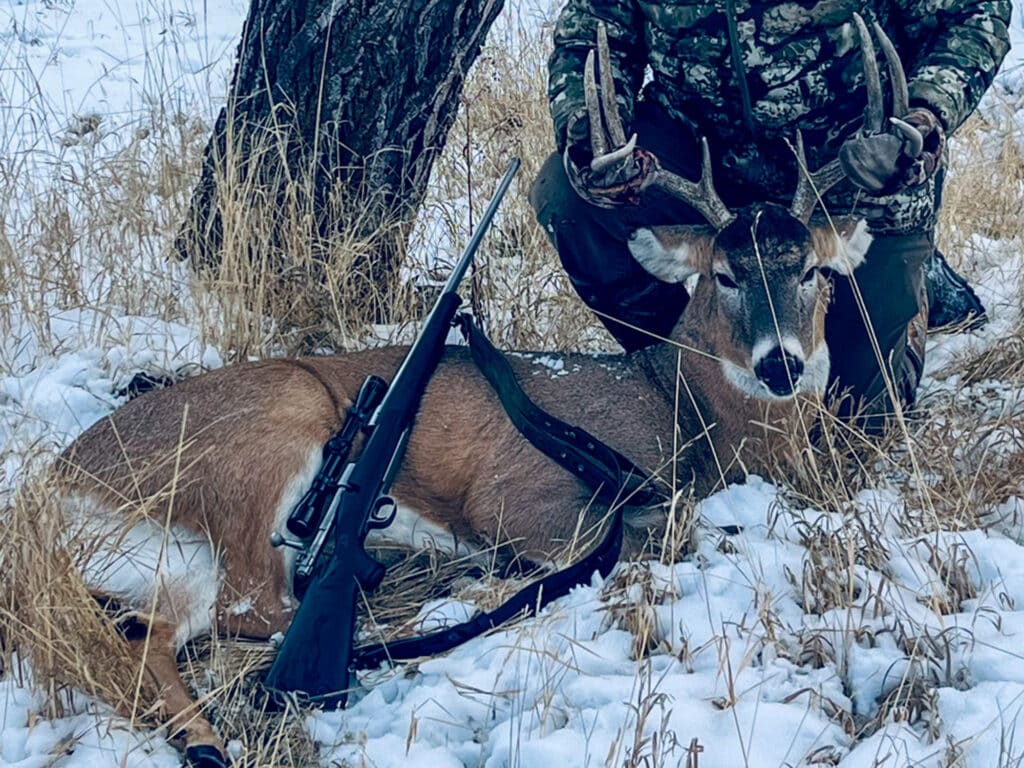 A person in camouflage kneels behind a deer with antlers lying on snowy ground at Rock Creek Meadows, holding its head up. A rifle with a scope rests on the deer’s side. Dry grass and a tree are visible in the background.