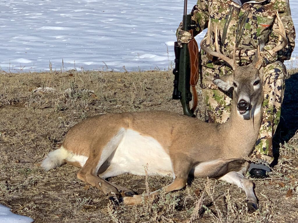 A person in camouflage holds a rifle and kneels next to a large deer with antlers lying on the ground in the snowy, grassy landscape of Rock Creek Meadows.