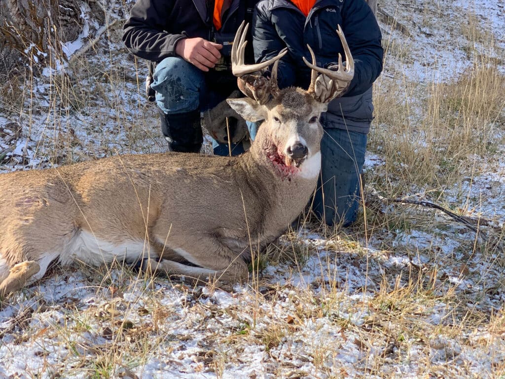 Two hunters kneel behind a large buck deer lying on snowy grass in Rock Creek Meadows. The deer has prominent antlers and a visible wound on its neck. The hunters’ faces are not shown.