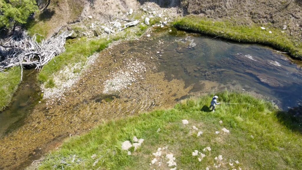 A person stands on the grassy bank at Rock Creek Meadows, fly fishing in a shallow, winding stream surrounded by rocks and green vegetation, viewed from above.