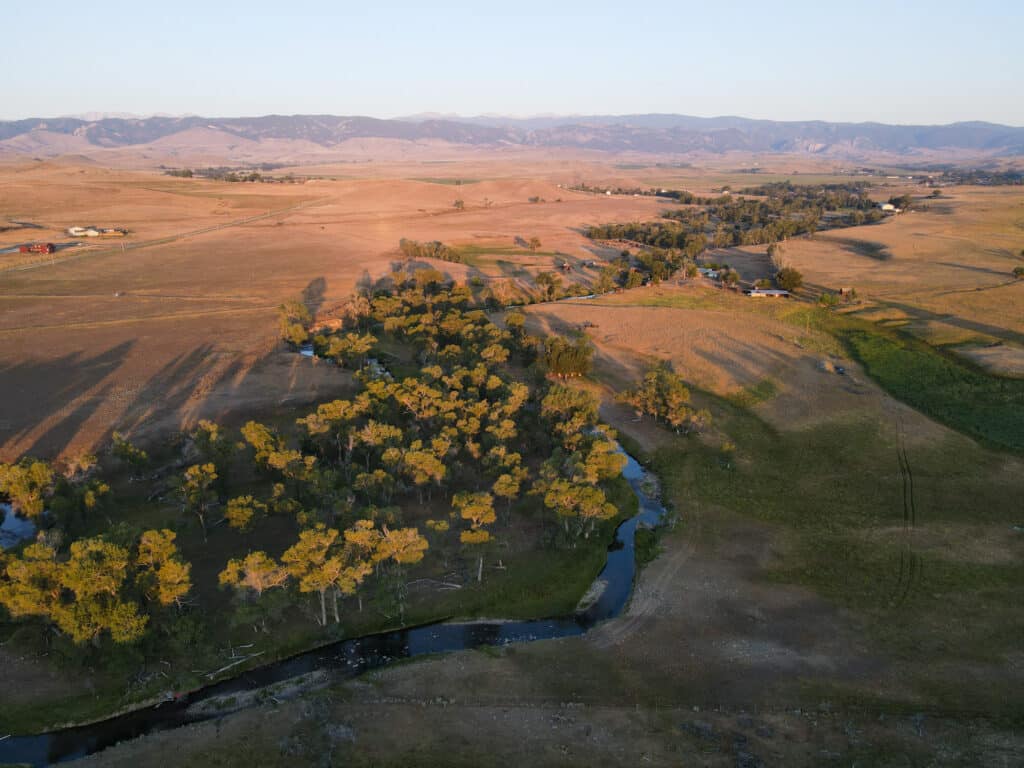 Aerial view of Rock Creek Meadows at sunset, with a winding creek, clusters of trees, open fields, scattered houses, and distant mountains under a clear sky.