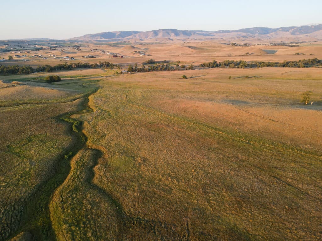 Aerial view of Rock Creek Meadows, a vast, dry grassy plain with patches of green, scattered houses, trees, and distant mountains under a clear sky.