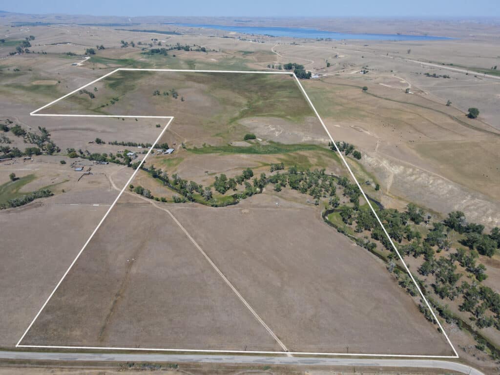 Aerial view of a large, dry, grassy landscape known as Rock Creek Meadows, with a white outline marking property boundaries; sparse trees and a creek run through the area, with water and hills in the distant background.