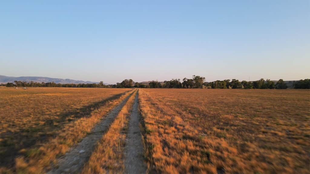 A dirt path runs through a wide, dry grassy field under a clear blue sky in Rock Creek Meadows, with distant trees and hills visible near the horizon.