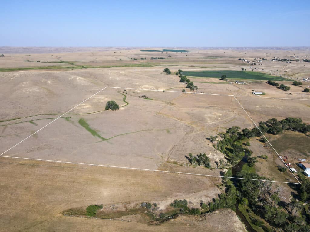 Aerial view of the expansive Rock Creek 40, a large, dry, open field bordered by a faint white outline, with some scattered trees, a winding Rock Creek, and distant farmland under a clear sky.
