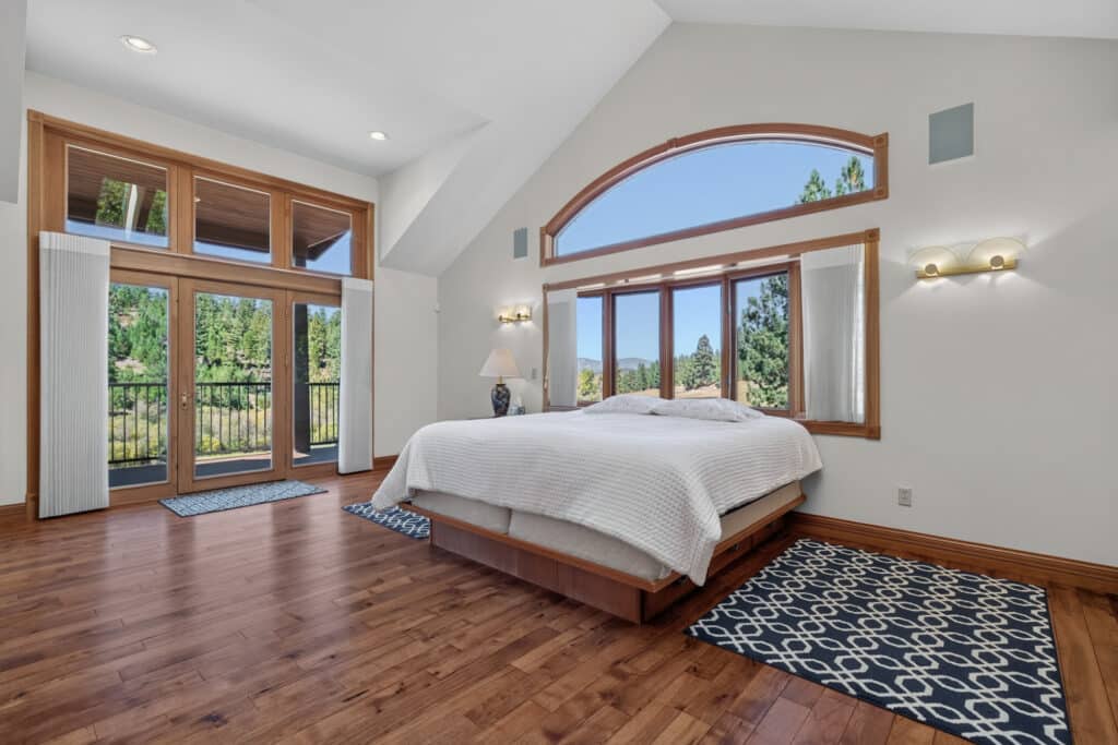 Bright bedroom with wood floors, large bed, and big windows showcasing trees and mountains. Glass doors open to a balcony in this Elkhorn sanctuary, where natural light and airy white walls create a serene retreat.