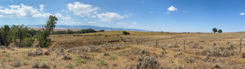 Wide panorama of a dry, grassy field with scattered trees and shrubs under a blue sky with clouds, distant hills, and mountains in the background. A rustic fence runs through the landscape, capturing the serene beauty of Rock Creek 40.