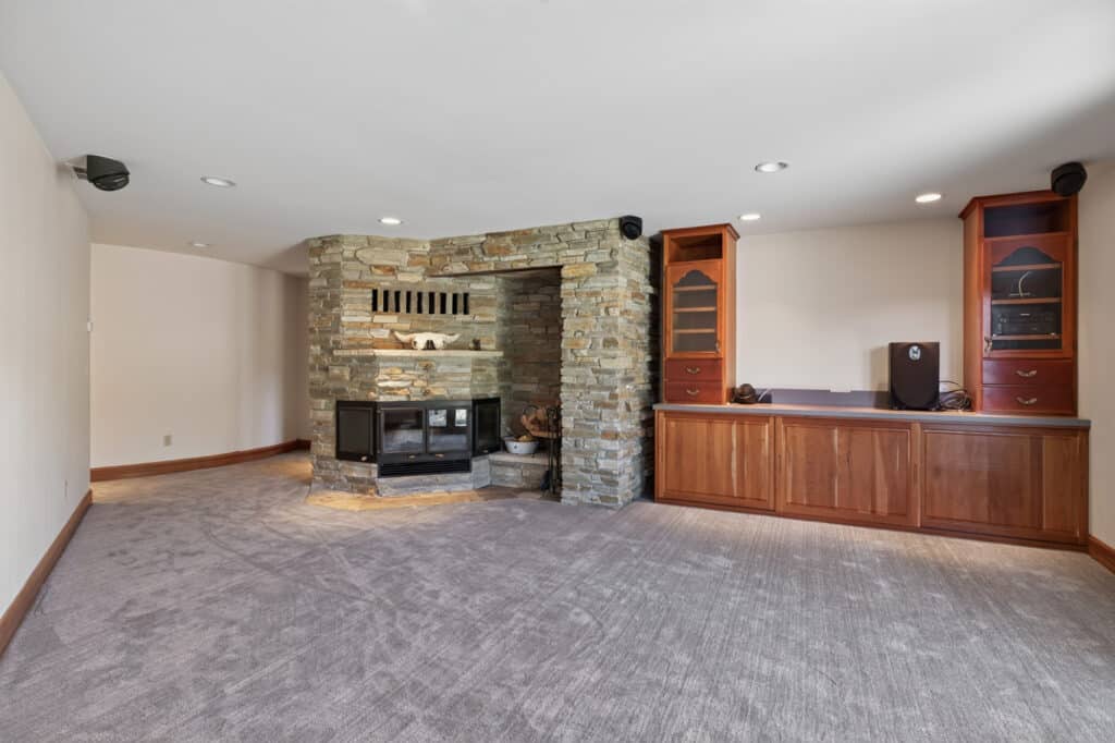 Spacious carpeted room with a stone fireplace, wood stove, built-in wood cabinets with glass doors, and recessed ceiling lights. This Elkhorn sanctuary features neutral walls and feels like a cozy basement or living space.