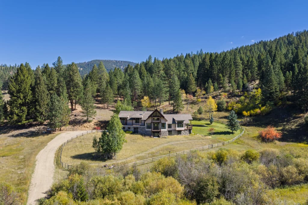 A large house with a wraparound porch sits among trees and hills at Elkhorn Sanctuary, surrounded by a fence and open grassy fields under a clear blue sky.