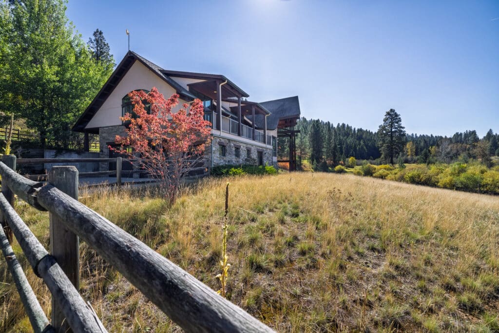 A modern house with large windows and a sloped roof sits in a grassy field at Elkhorn sanctuary, bordered by a wooden fence and surrounded by trees under a clear blue sky.