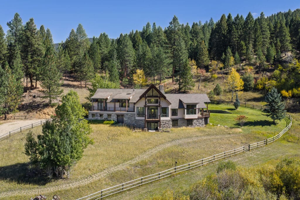 A large house with stone and wood accents sits on a grassy lot in the Elkhorn sanctuary, surrounded by a wooden fence, trees, and forested hills under a clear blue sky.