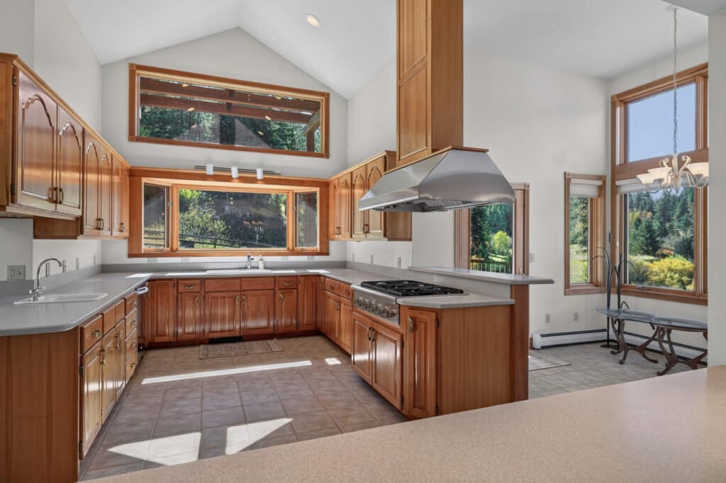 Spacious kitchen with wood cabinets, stainless steel range hood, large windows, ample counter space, tiled floor, and a view of trees and the Elkhorn sanctuary garden outside.