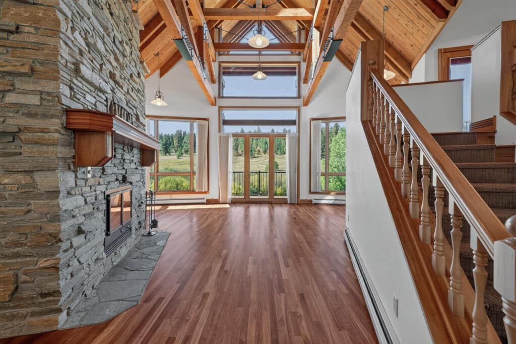 Spacious living room with wood floors, stone fireplace, vaulted wood-beam ceiling, large windows providing scenic views, and a wooden staircase on the right—your Elkhorn sanctuary awaits.