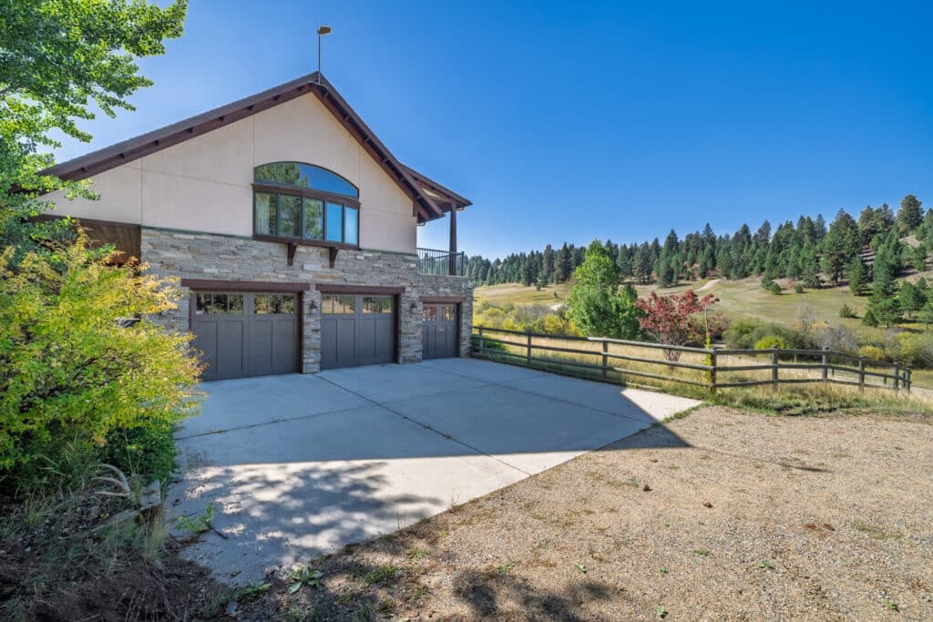 Two-car garage home with stone and wood exterior, large window, and balcony, nestled in the Elkhorn sanctuary among trees and hills under a clear blue sky. Concrete driveway and wooden fence in front.