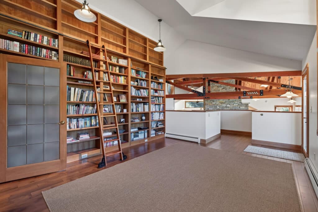 A spacious library room with floor-to-ceiling bookshelves, a ladder, wooden floors, and exposed beam ceilings offers an Elkhorn sanctuary feel. Large windows brighten doors on the left and an open view to the right.