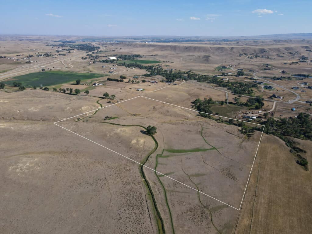 Aerial view of a dry, rural landscape with rolling hills, sparse greenery, scattered houses, and a white boundary outlining the expansive Rock Creek 40 plot of land.