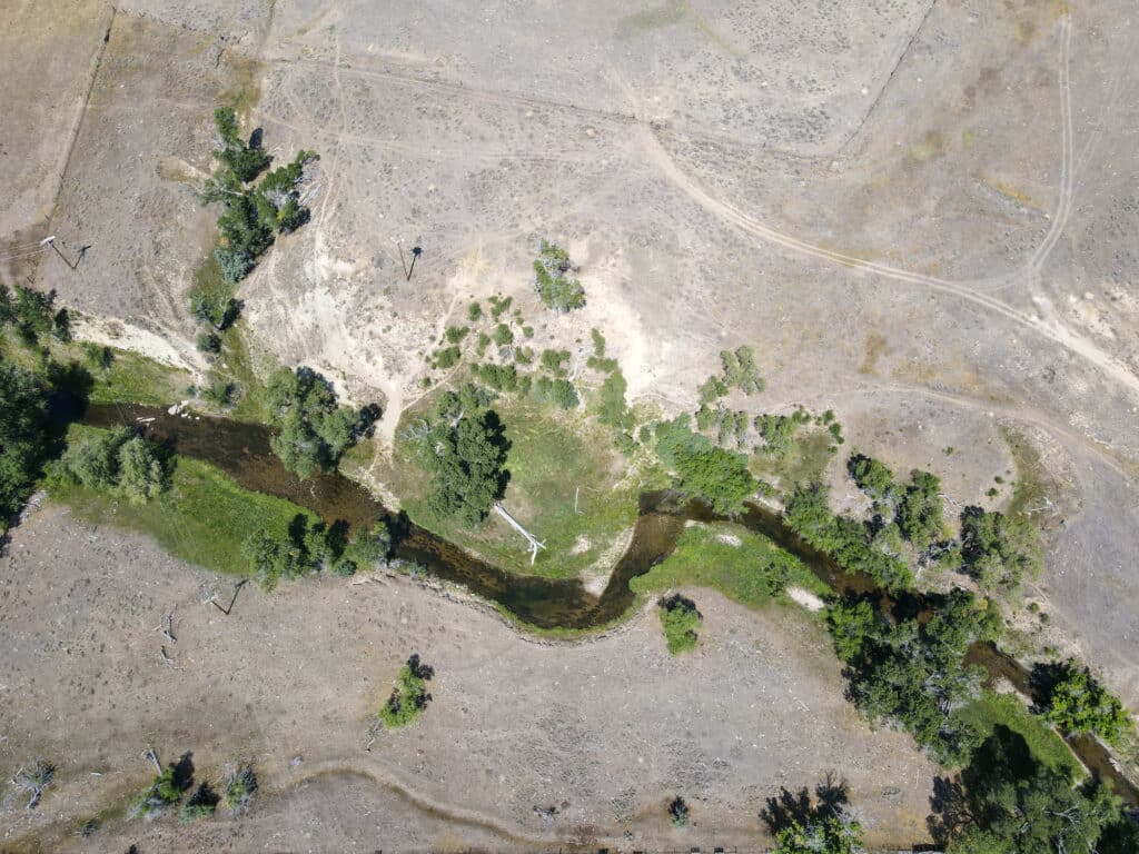 Aerial view of Rock Creek 40, a small, winding creek bordered by green trees and surrounded by dry, brown grassland with faint paths and patches of bare soil.