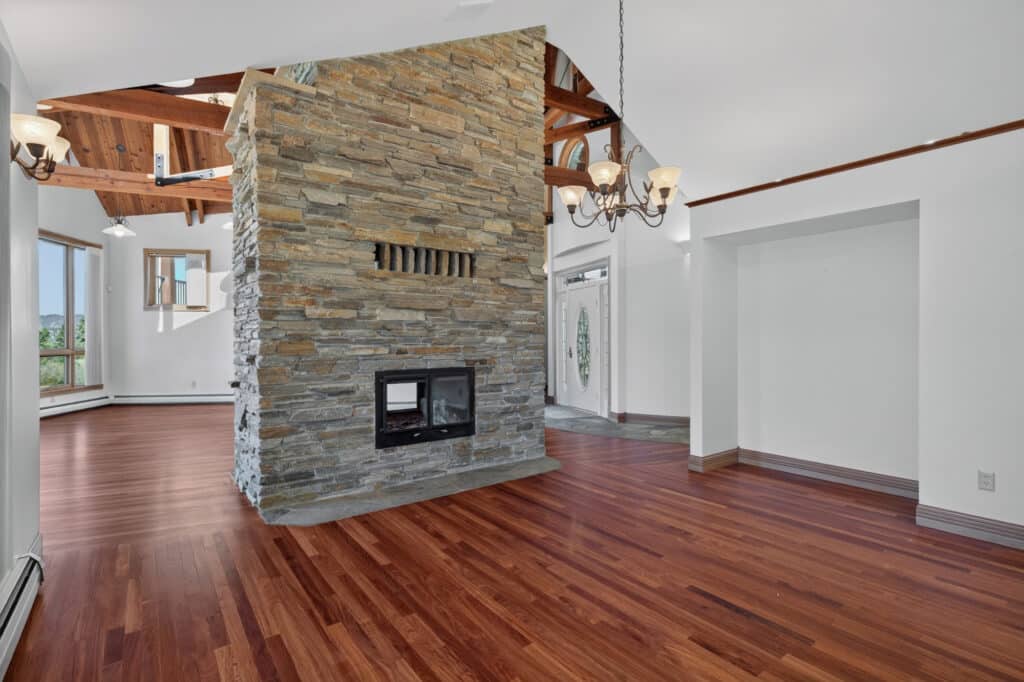Open living space with rich wood floors, a central double-sided stone fireplace, large windows, chandelier, exposed wood beams, and an arched ceiling—your modern Elkhorn sanctuary for warmth and inviting comfort.