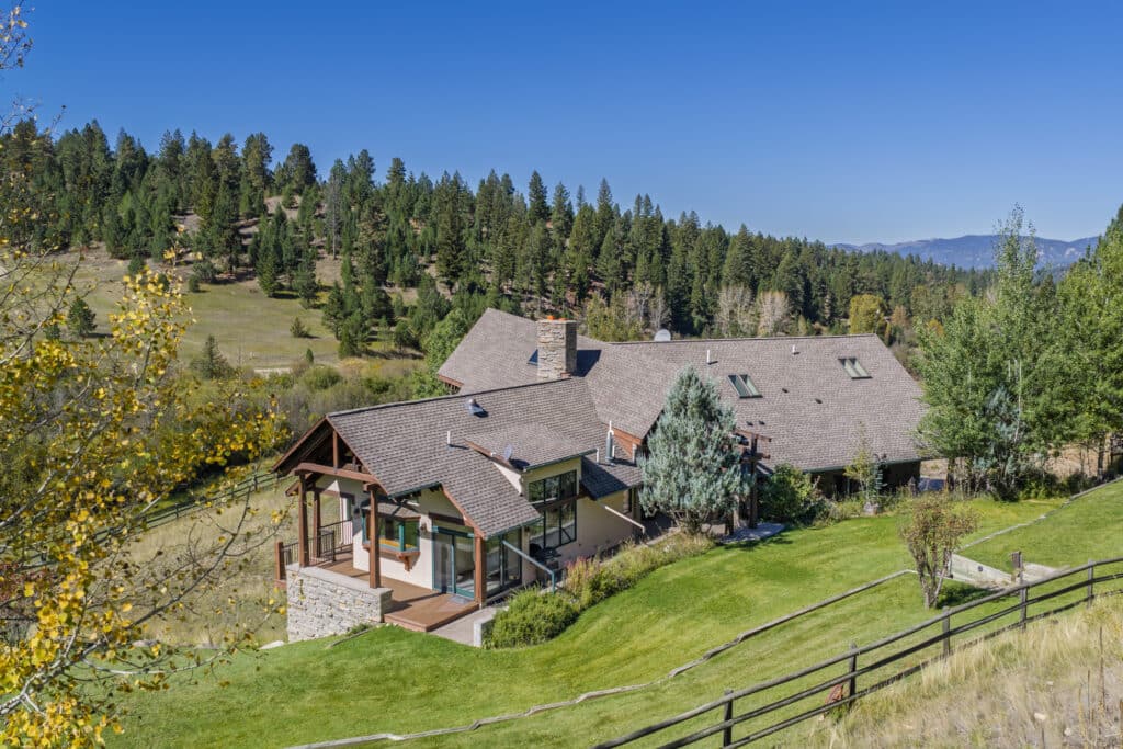 Large rustic home with stone and wood accents, set on a grassy hillside at Elkhorn sanctuary, surrounded by trees and mountains under a clear blue sky.