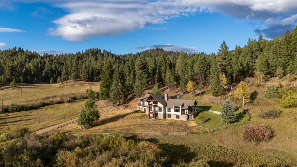 A large house with wood and stone accents sits in a grassy clearing, surrounded by trees and forested hills—an Elkhorn sanctuary under a partly cloudy blue sky.