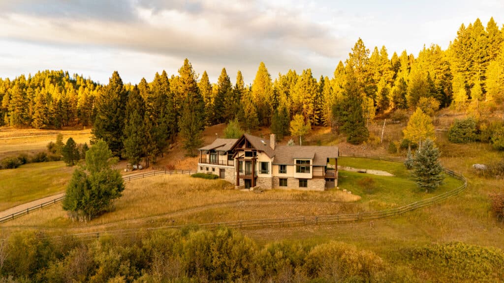 A large Elkhorn house with stone and wood exterior sits in a grassy field, surrounded by a wooden fence and dense pine trees, bathed in warm sunlight under a partly cloudy sky—a true sanctuary.