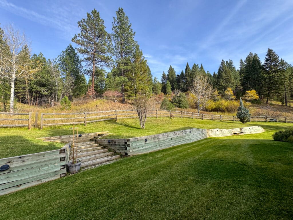 A terraced grassy lawn at the Elkhorn nature reserve, with wood retaining walls and stairs leading up to a wooden fence and a pine forest.